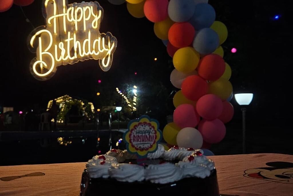 A birthday cake with a 'Happy Birthday' topper, surrounded by balloons and a neon sign in the background.