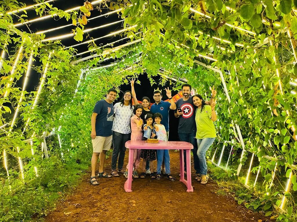 A group of people gathered under a vine-covered structure with a pink table in the center.