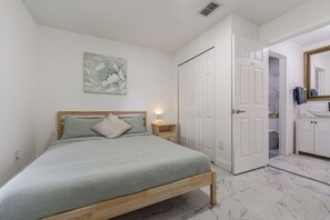 A bedroom with a bed, a nightstand, a wall-mounted lamp, a door, and a bathroom visible through an open door. - Poolside Fun 8 Mi to Disney! Family Getaway (Kissimmee)