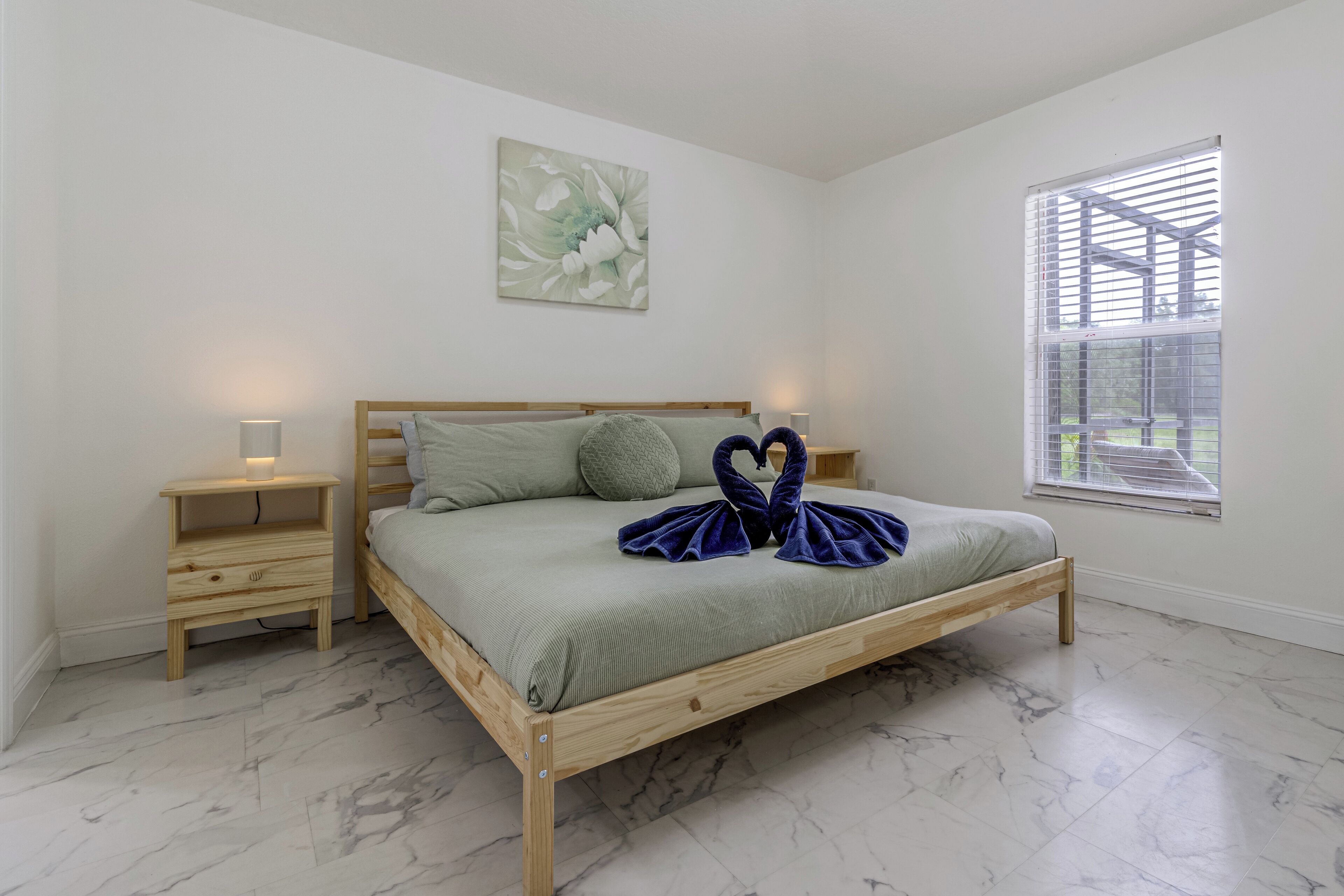 Een slaapkamer met een houten bed, een nachtkastje, een lamp, een raam met jaloezieën en een schilderij aan de muur.