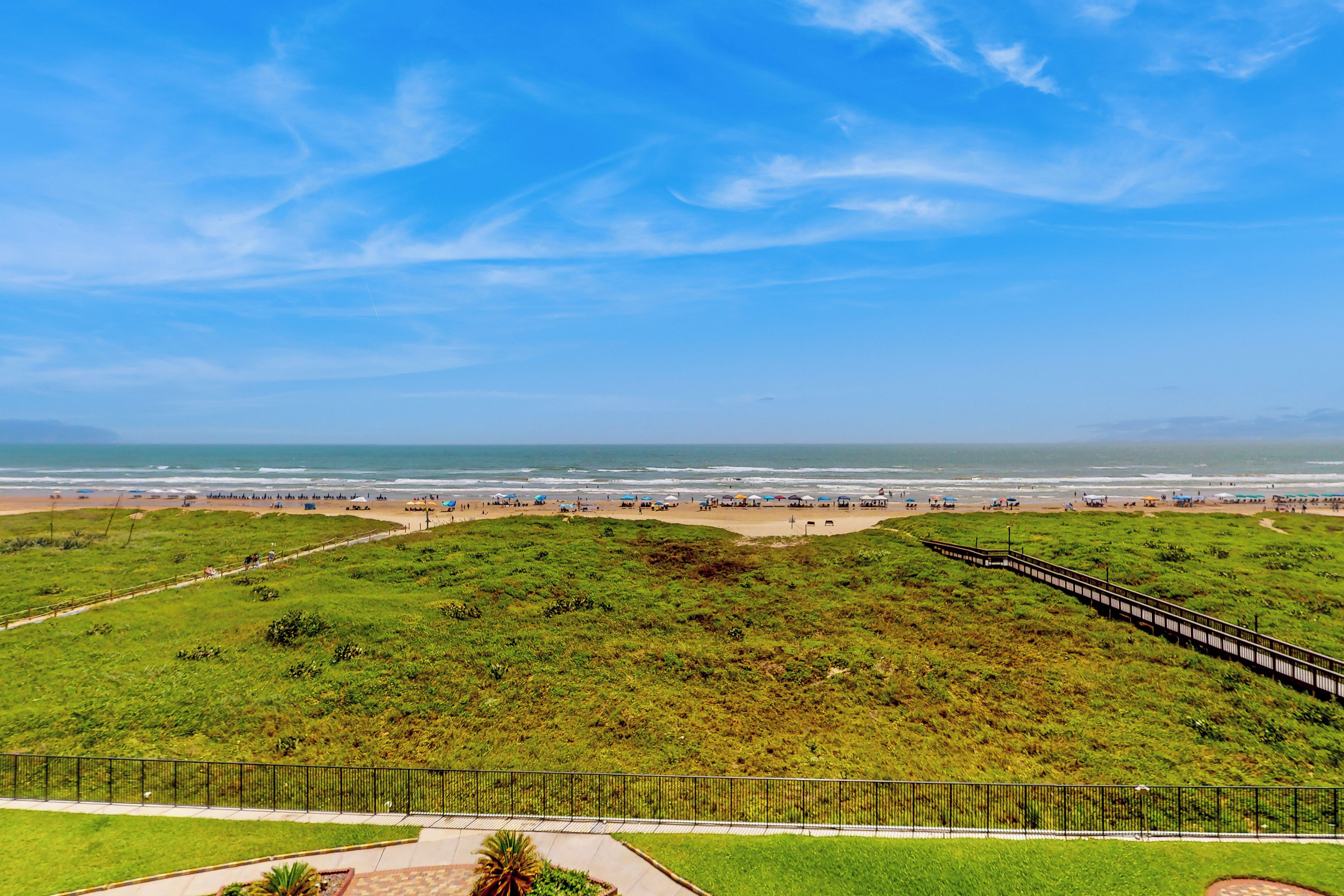 A coastal landscape with a grassy area, a walking path, and a beach with umbrellas.
