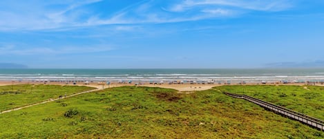 Paisaje costero con una zona de césped, un sendero y una playa con sombrillas.