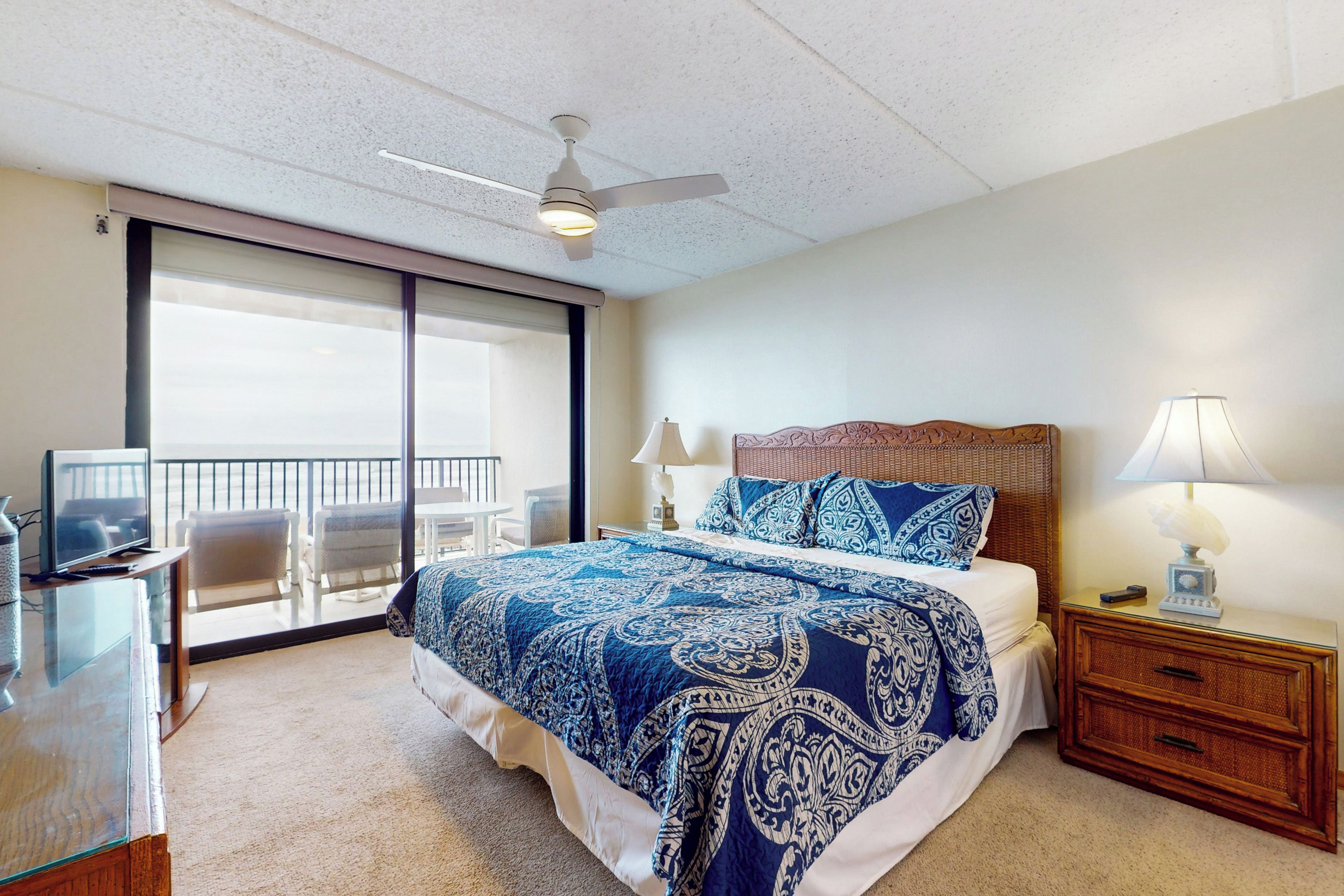 A bedroom with a large bed, a ceiling fan, a TV on a stand, a nightstand with a lamp, and a balcony with chairs.