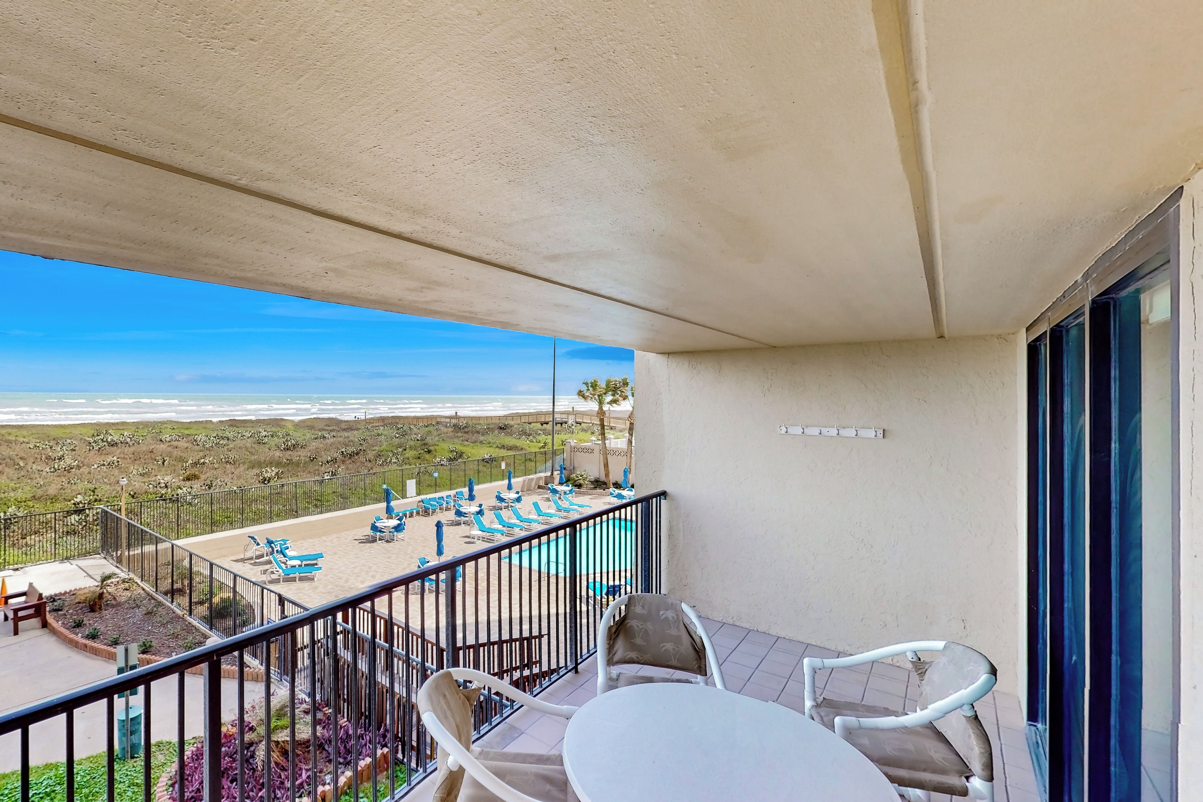 A balcony with a view of the beach, a pool area with lounge chairs, and a table with chairs.