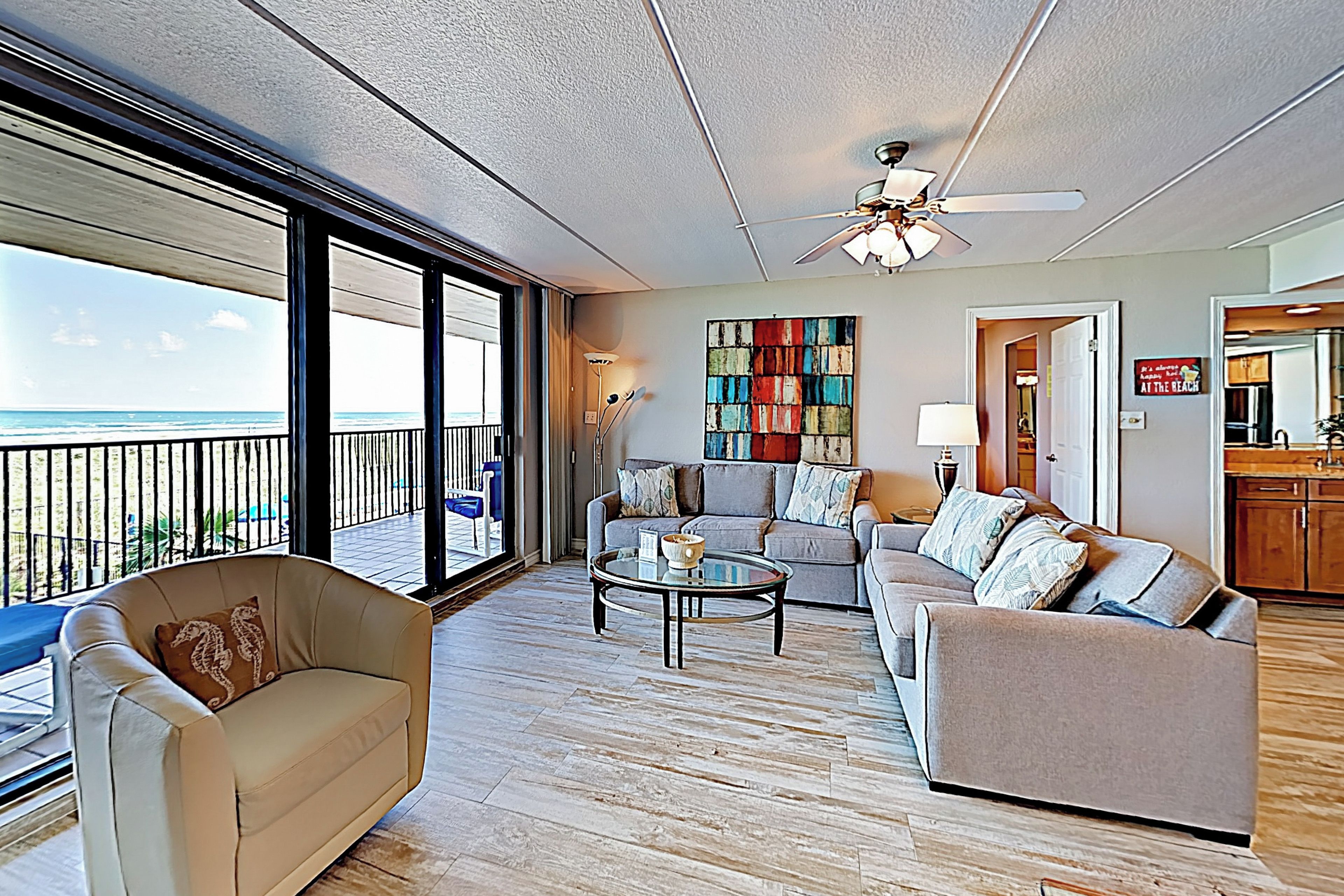 A spacious living area with a balcony overlooking the ocean, a ceiling fan, and a colorful abstract painting on the wall.