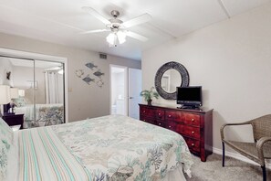 Ein Schlafzimmer mit Bett, Deckenventilator, Schminktisch mit Fernseher, Stuhl und Spiegel.