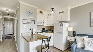 A kitchen with white cabinets, a granite countertop, a stove, a refrigerator, and a dining area with a chair. - Poolside Vibes, Beach Strolls! Galveston Getaway (Galveston)