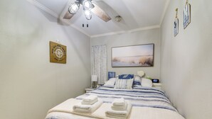 A bedroom with a bed, a ceiling fan, a painting, a clock, and a wall decoration. - Poolside Vibes, Beach Strolls! Galveston Getaway (Galveston)