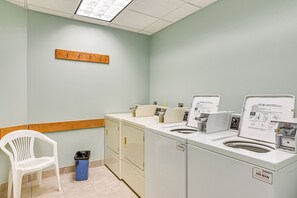 A laundry room with two washers and dryers, a white plastic chair, and a wooden coat rack. - Poolside Vibes, Beach Strolls! Galveston Getaway (Galveston)