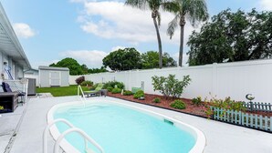 Ein Swimmingpool mit weißer Liegefläche, umgeben von einem weißen Zaun, mit einer kleinen Gartenanlage und einer Palme im Hintergrund.