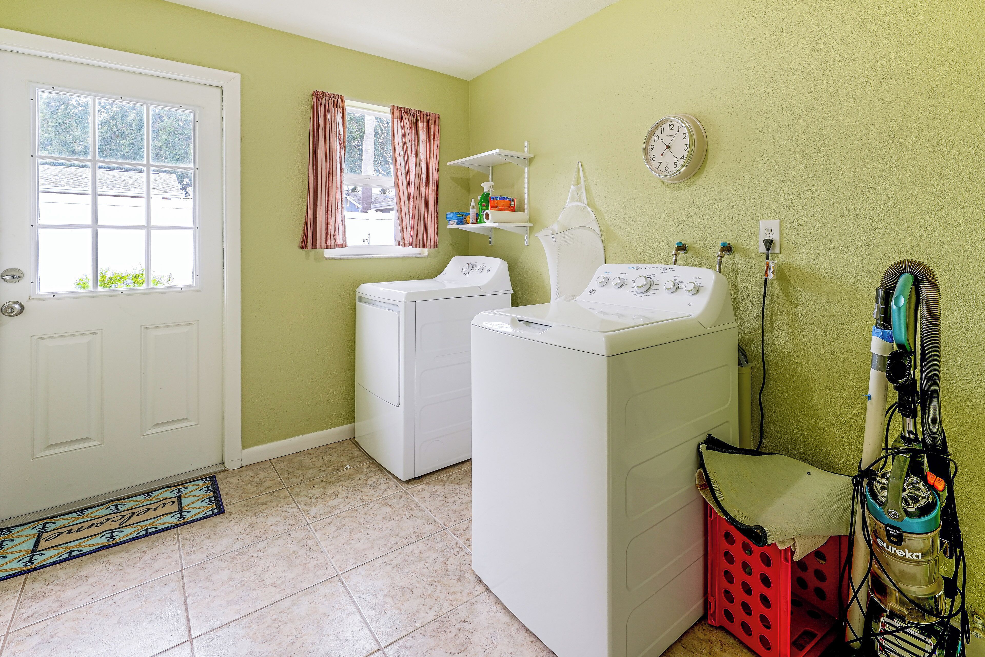 Une buanderie équipée d’un lave-linge et d’un sèche-linge à chargement frontal, d’une fenêtre avec des rideaux, d’une horloge au mur et d’un aspirateur.