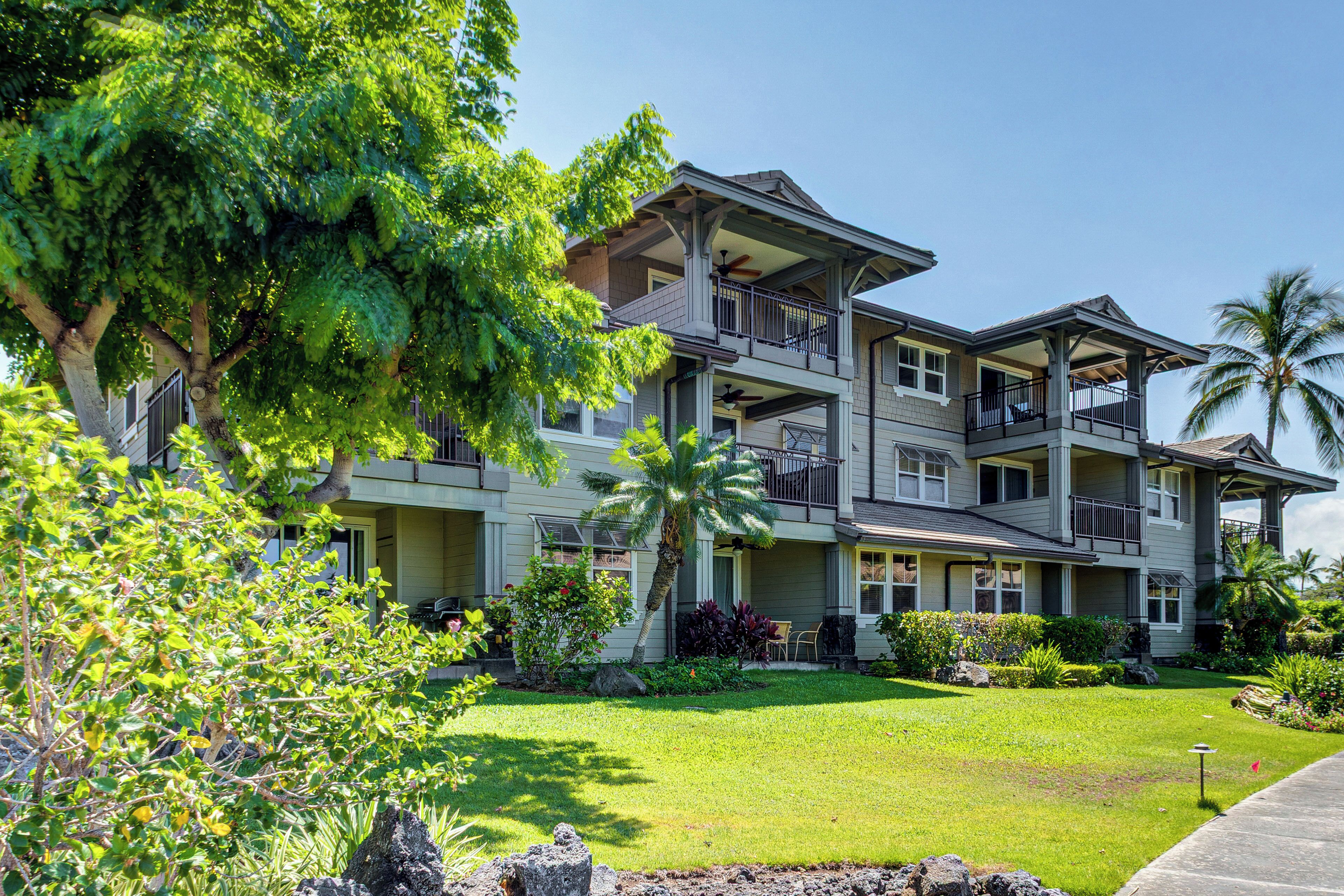 A multi-story hotel with a balcony on each floor, surrounded by lush greenery and palm trees.