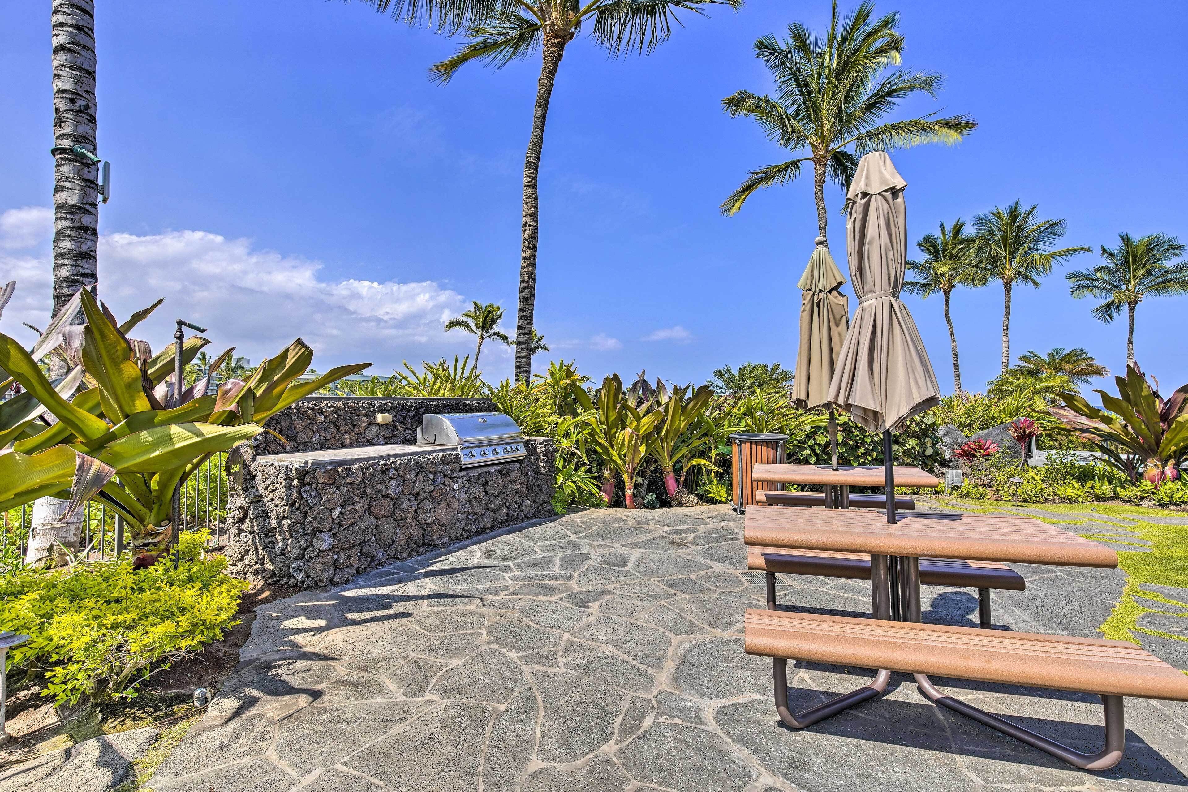 An outdoor patio area with stone flooring, a barbecue grill, picnic tables, and palm trees.