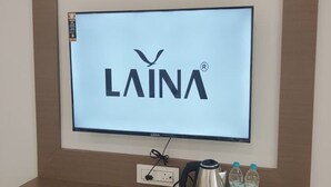 A television mounted on a wall with the brand 'LAINA' displayed, a power outlet, a remote control, a kettle, and a tray with bottles on a wooden console below. - Jayesh Lodging (Shirur)