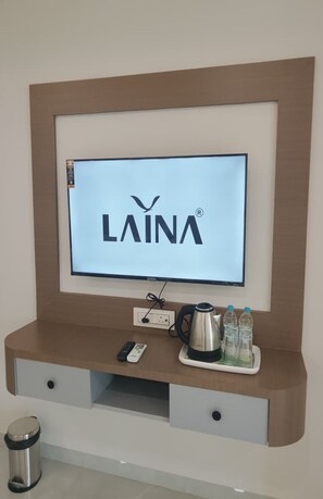 Ein an der Wand befestigter Fernseher mit dem Schriftzug 'LAINA', eine Steckdose, eine Fernbedienung, ein Wasserkocher und ein Tablett mit Flaschen auf einer Holzkonsole.