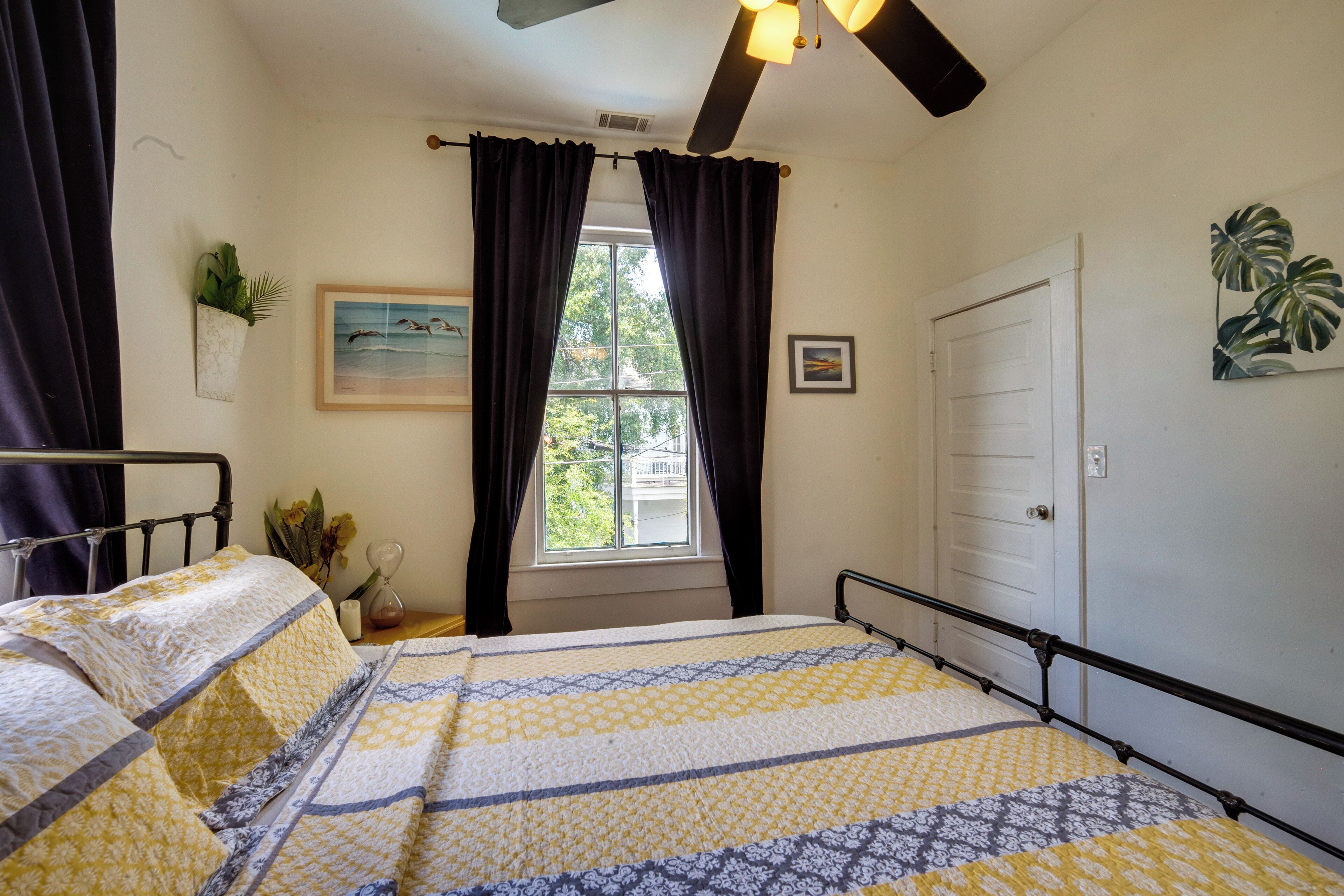 Une chambre à coucher avec un lit, une fenêtre avec des rideaux, un ventilateur de plafond, une porte et une plante fixée au mur.