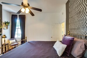 Ein Schlafzimmer mit einem Bett, einem Deckenventilator, einem Fenster mit Vorhängen, einem Sessel, einem Nachttisch mit Lampe und einem dekorativen Wandpaneel.