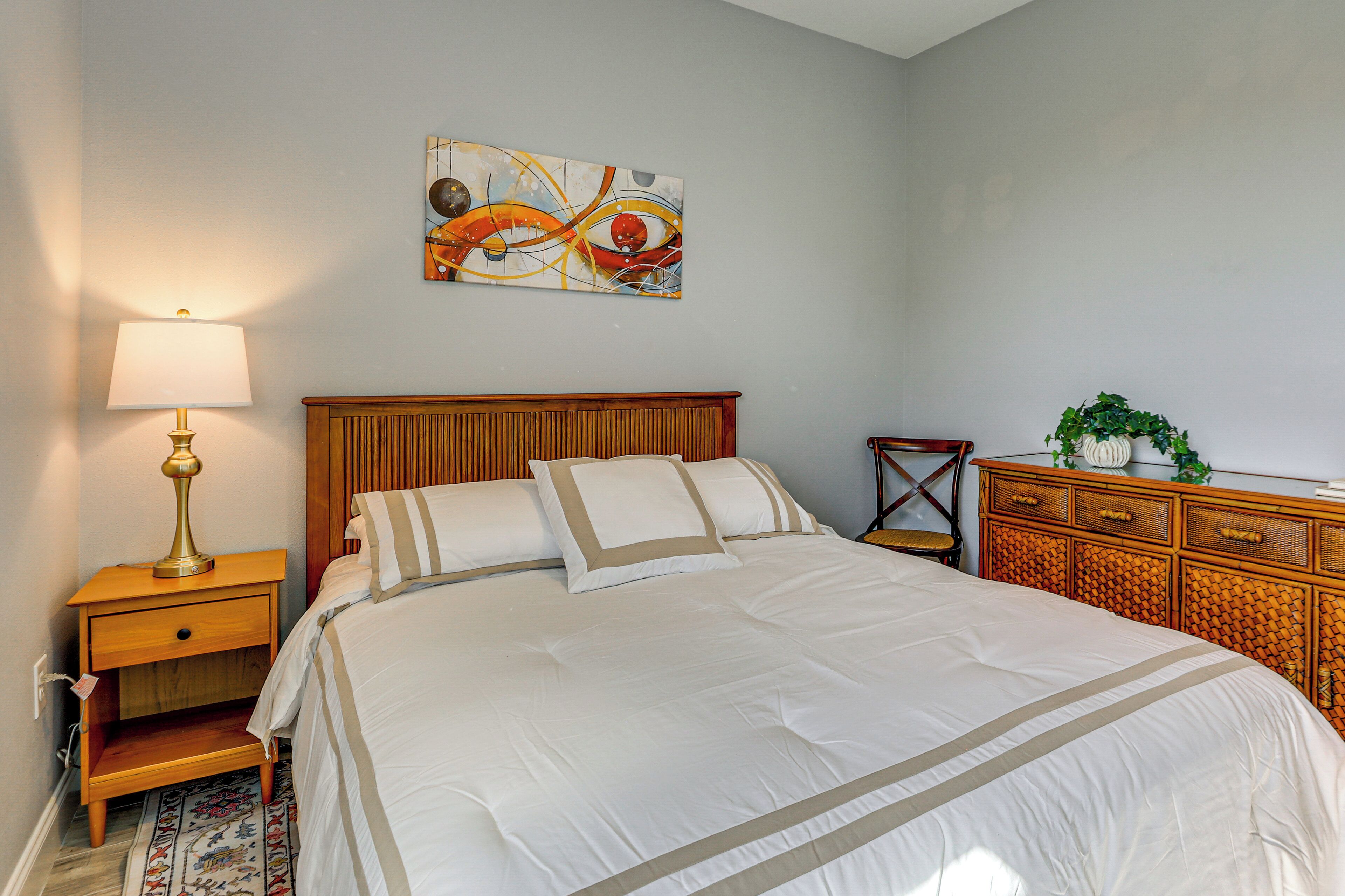 A neatly made bed with a wooden headboard, a bedside table with a lamp, a decorative wall piece, and a wicker side table with a plant.