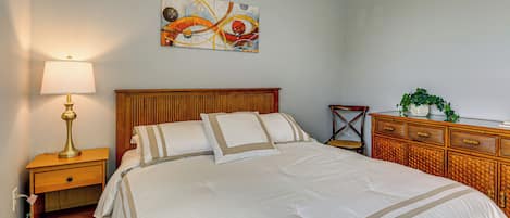 A neatly made bed with a wooden headboard, a bedside table with a lamp, a decorative wall piece, and a wicker side table with a plant.