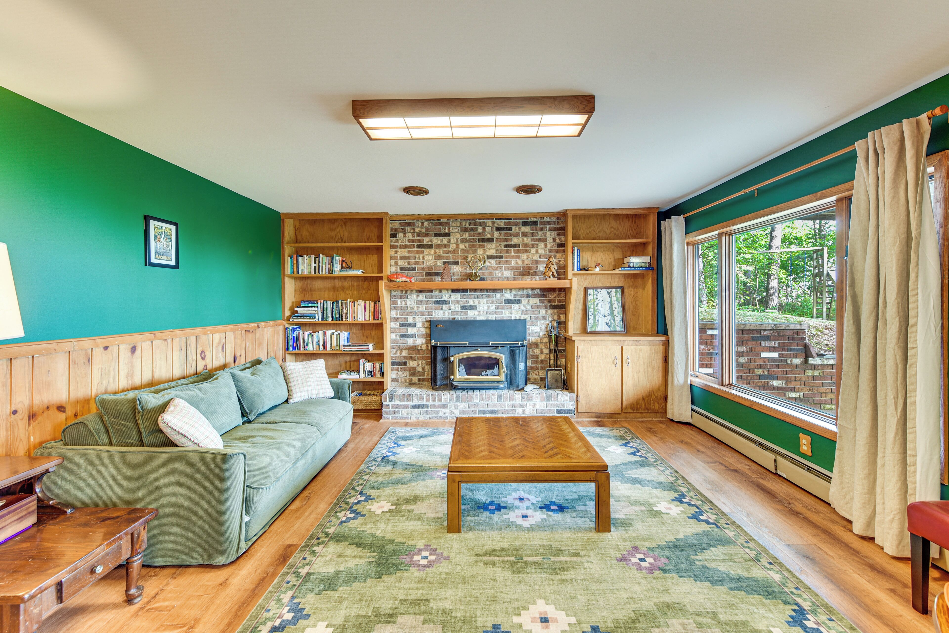 A cozy living room with a green wall, wooden furniture, a fireplace, and a large window with curtains.