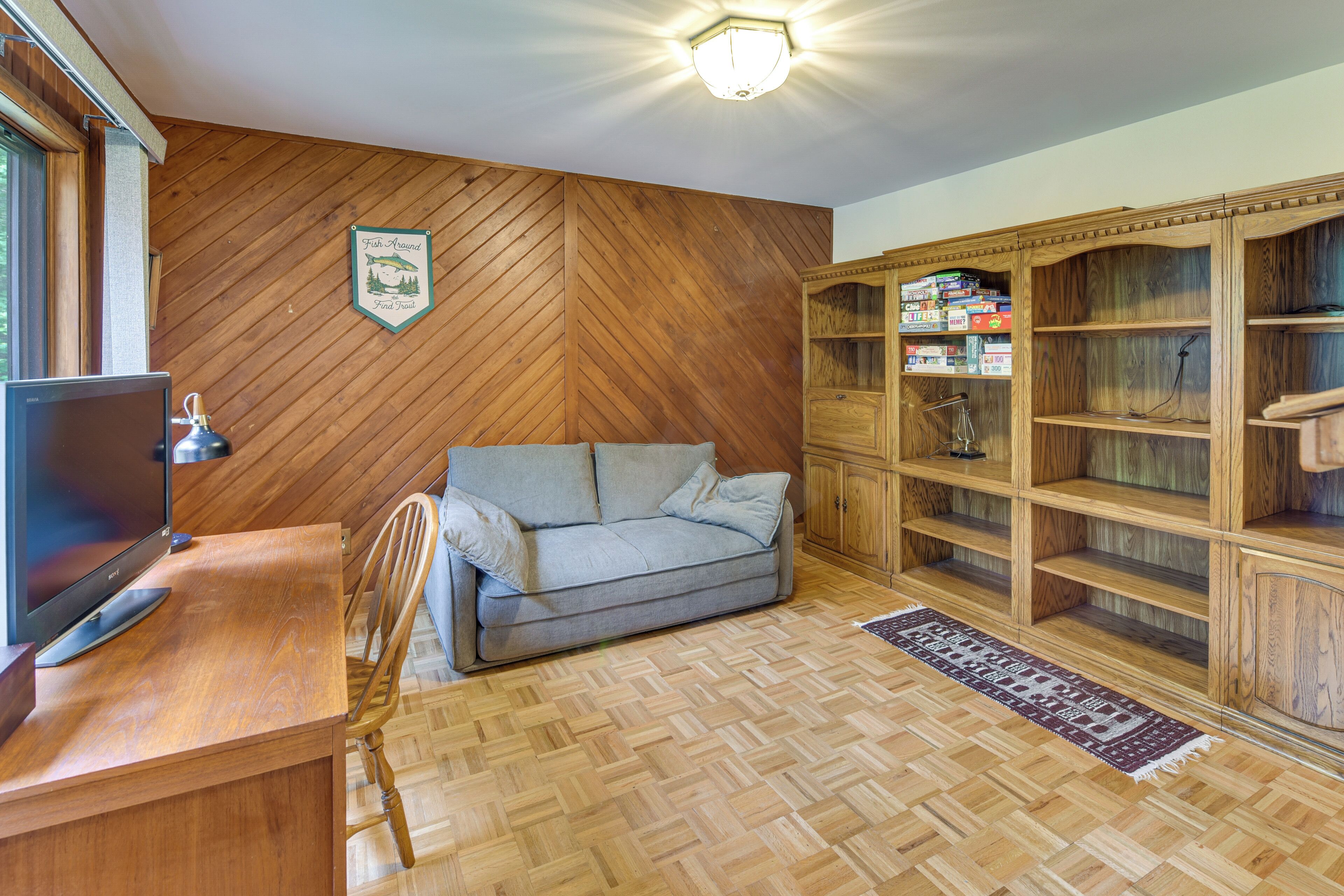 A room with a herringbone wooden floor, a grey sofa, a wooden cabinet with board games, and a television on a wooden desk.