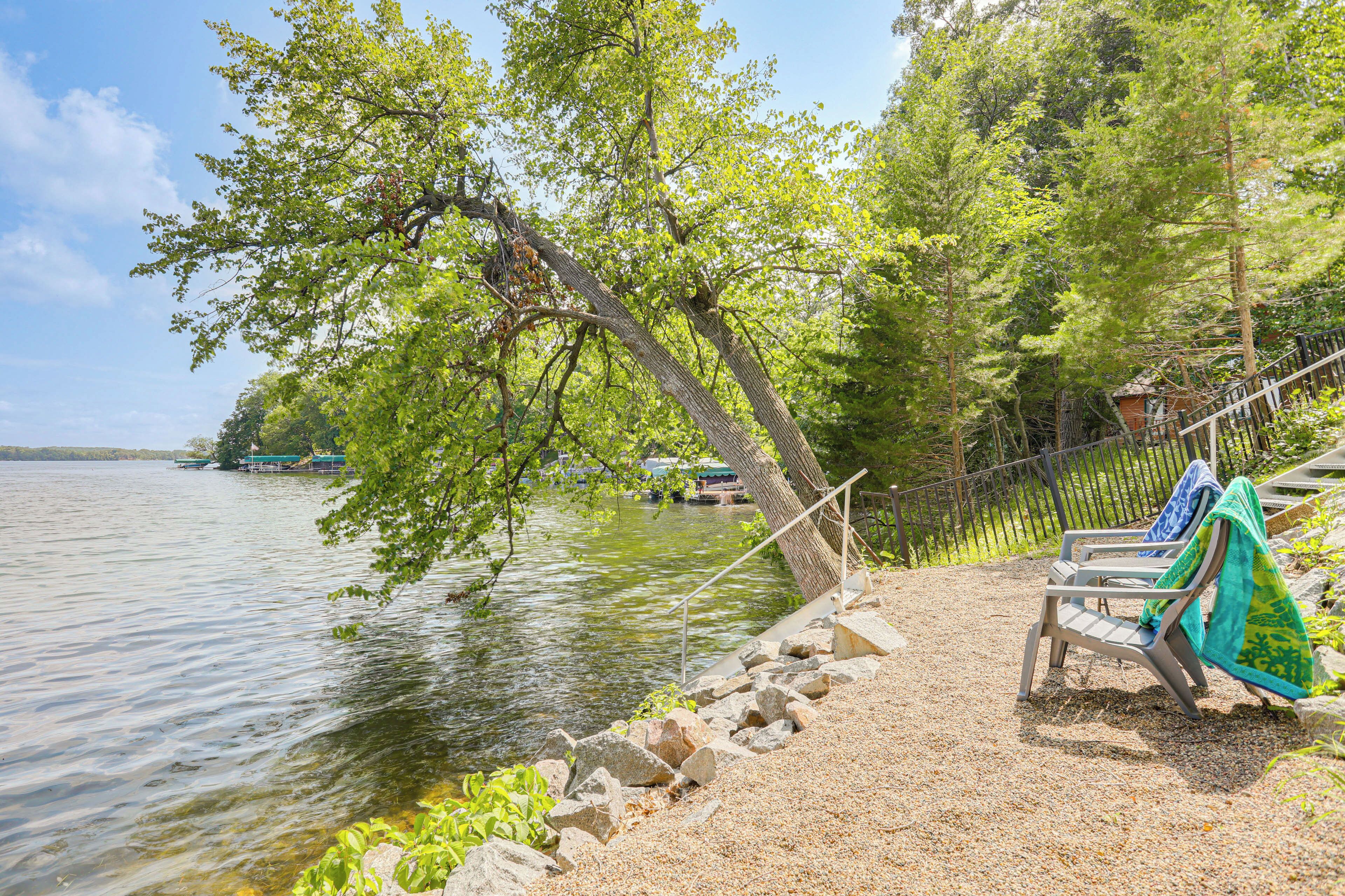 A lakeside seating area with a bench and a table, surrounded by trees and a rocky shoreline.