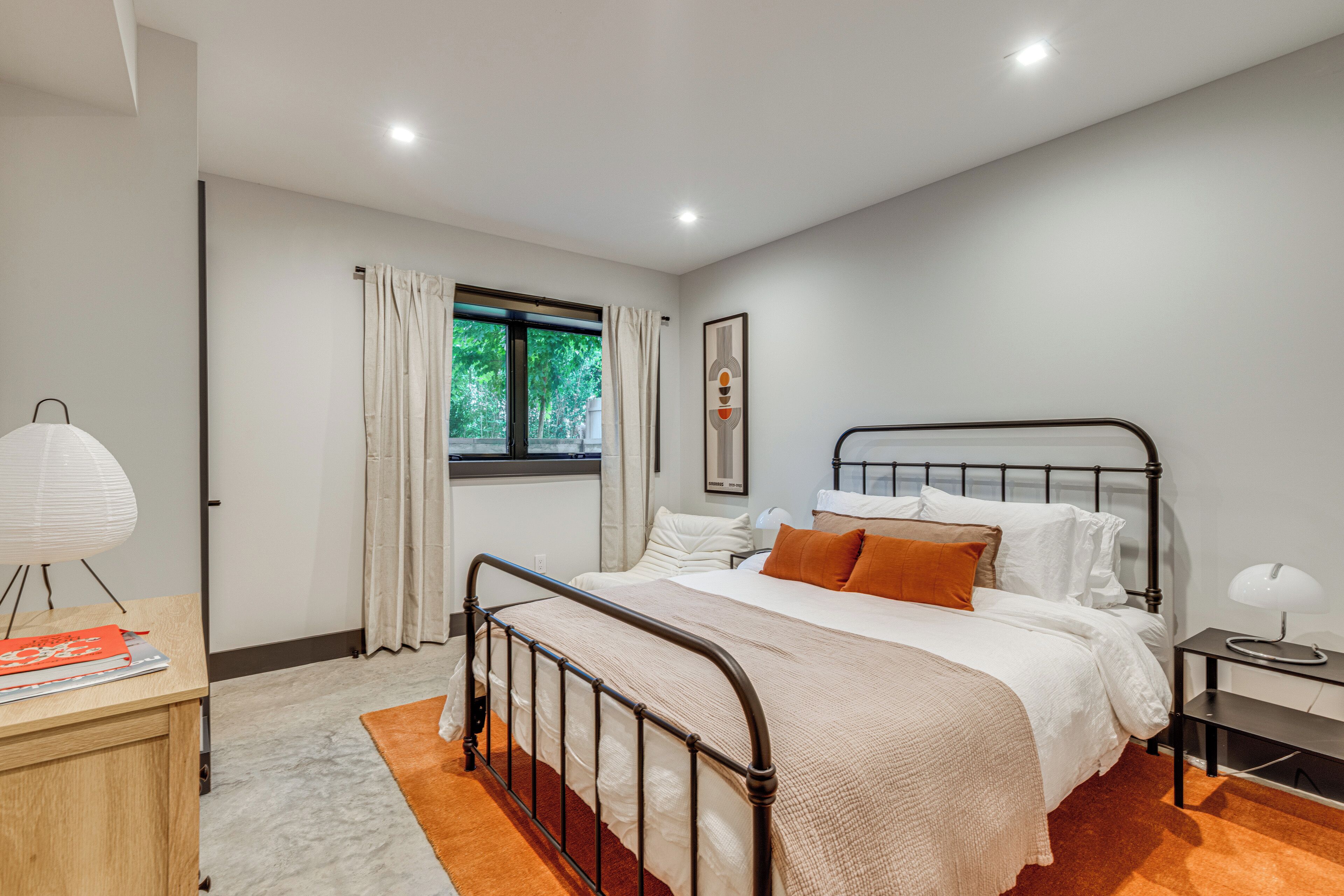 A bedroom with a bed, a nightstand, a lamp, a window with curtains, and a wall-mounted picture.