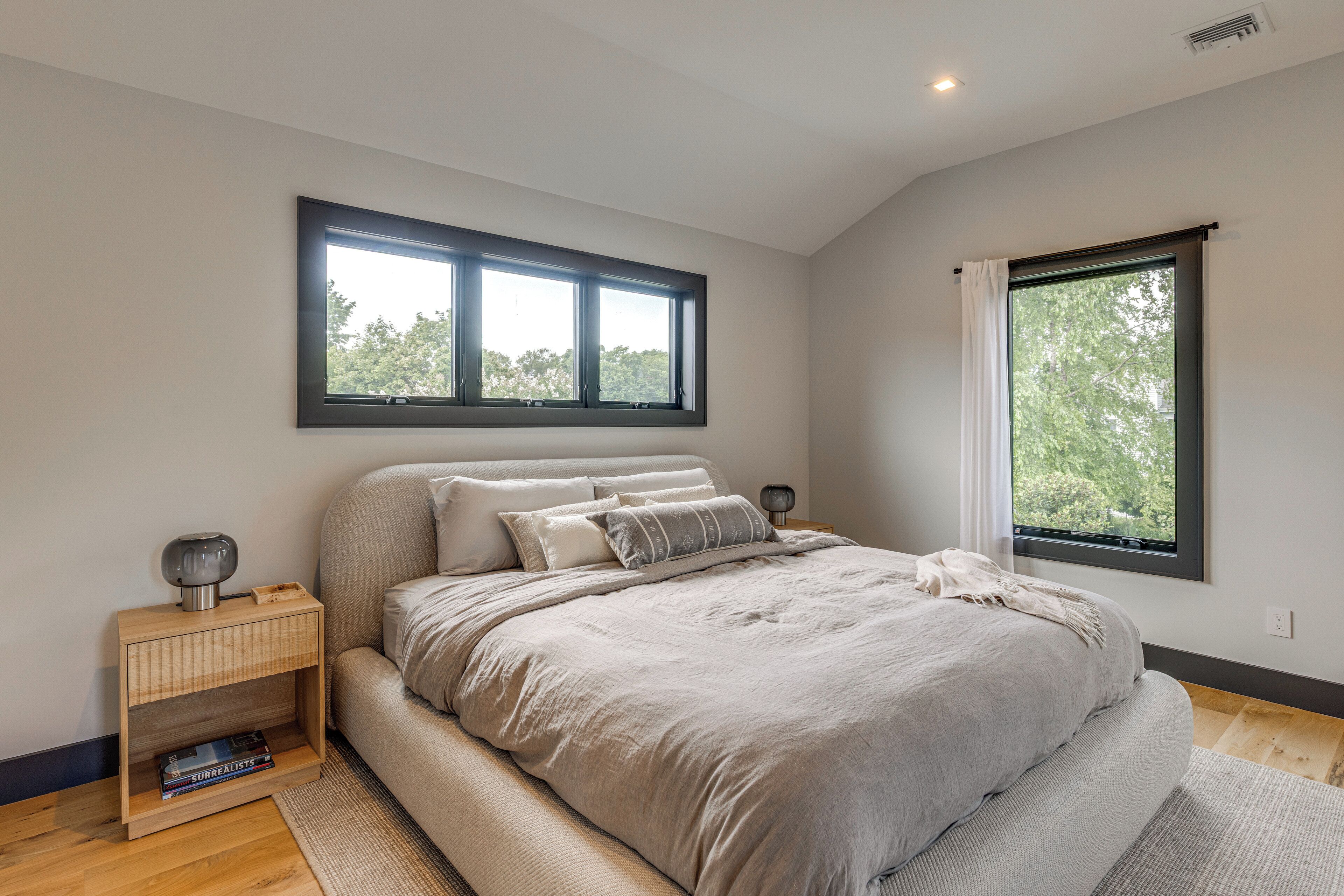 A bedroom with a large bed, a bedside table, a nightstand, a window with curtains, and a view of trees.