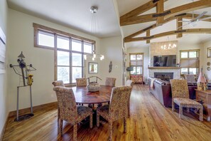 A spacious dining area with a wooden table and patterned chairs, a kitchen with a stone fireplace, and a large window providing natural light.