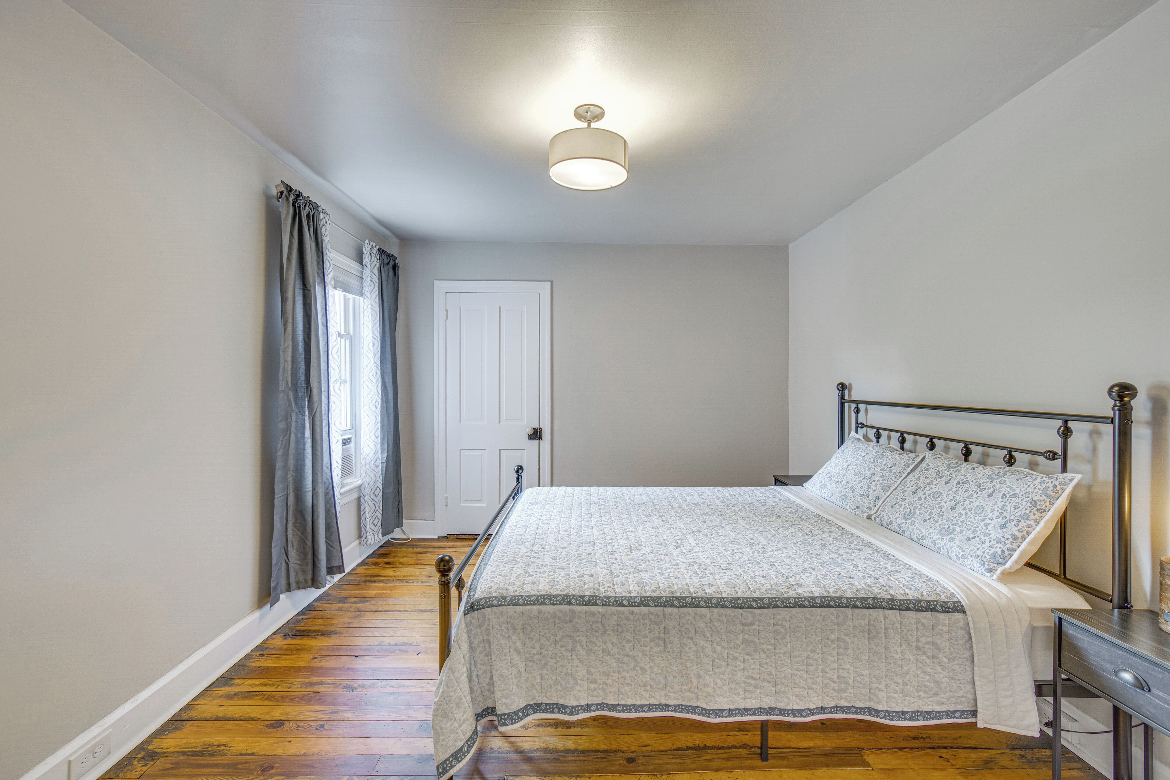 A bedroom with a bed, a bedside table, a nightstand, a window with curtains, and a door.