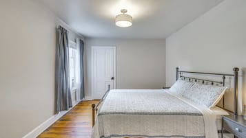 A bedroom with a bed, a bedside table, a nightstand, a window with curtains, and a door.