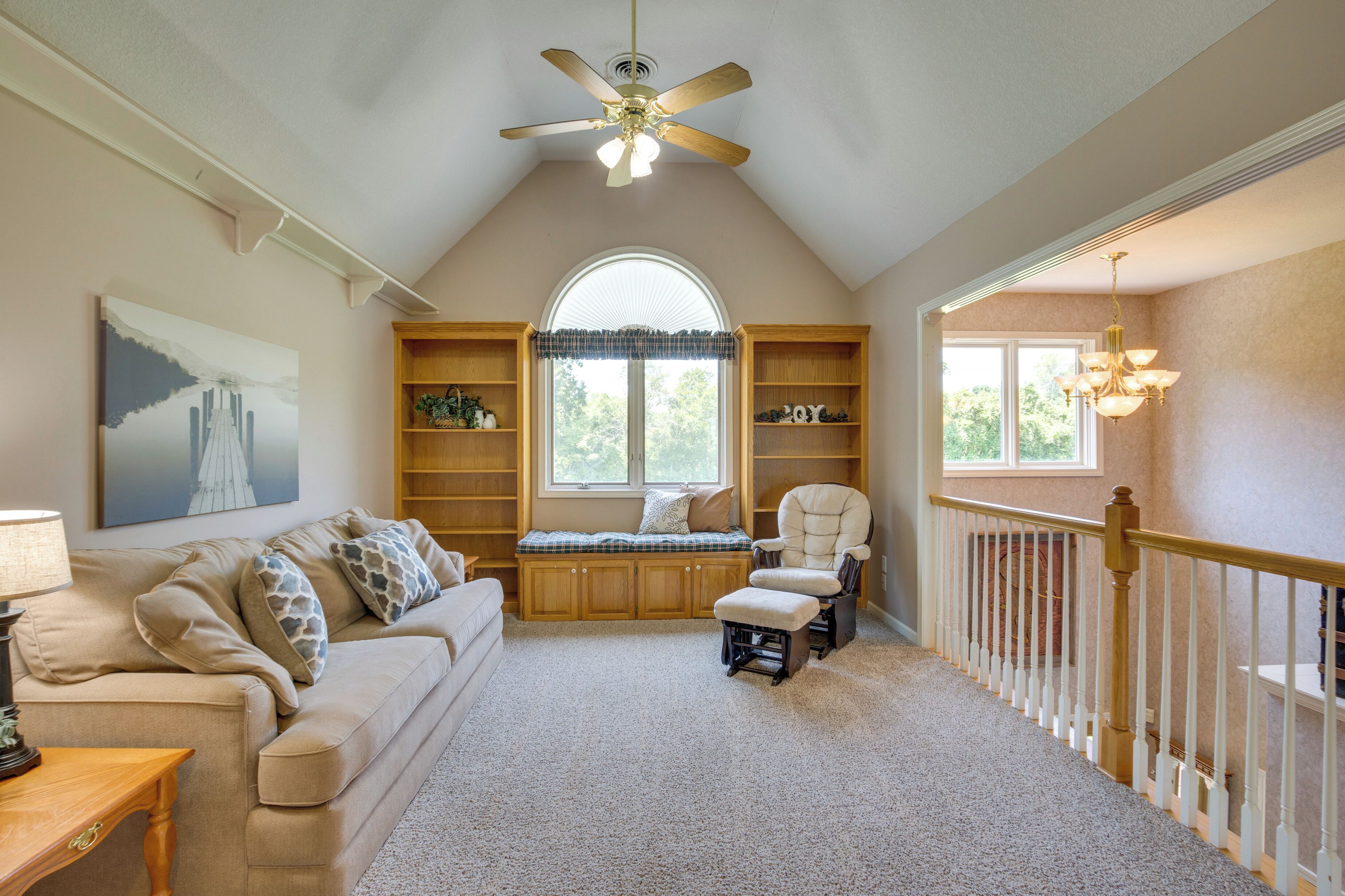 A cozy room with a high ceiling, a ceiling fan, a large window, a cushioned bench, a chair, and a staircase with a railing.