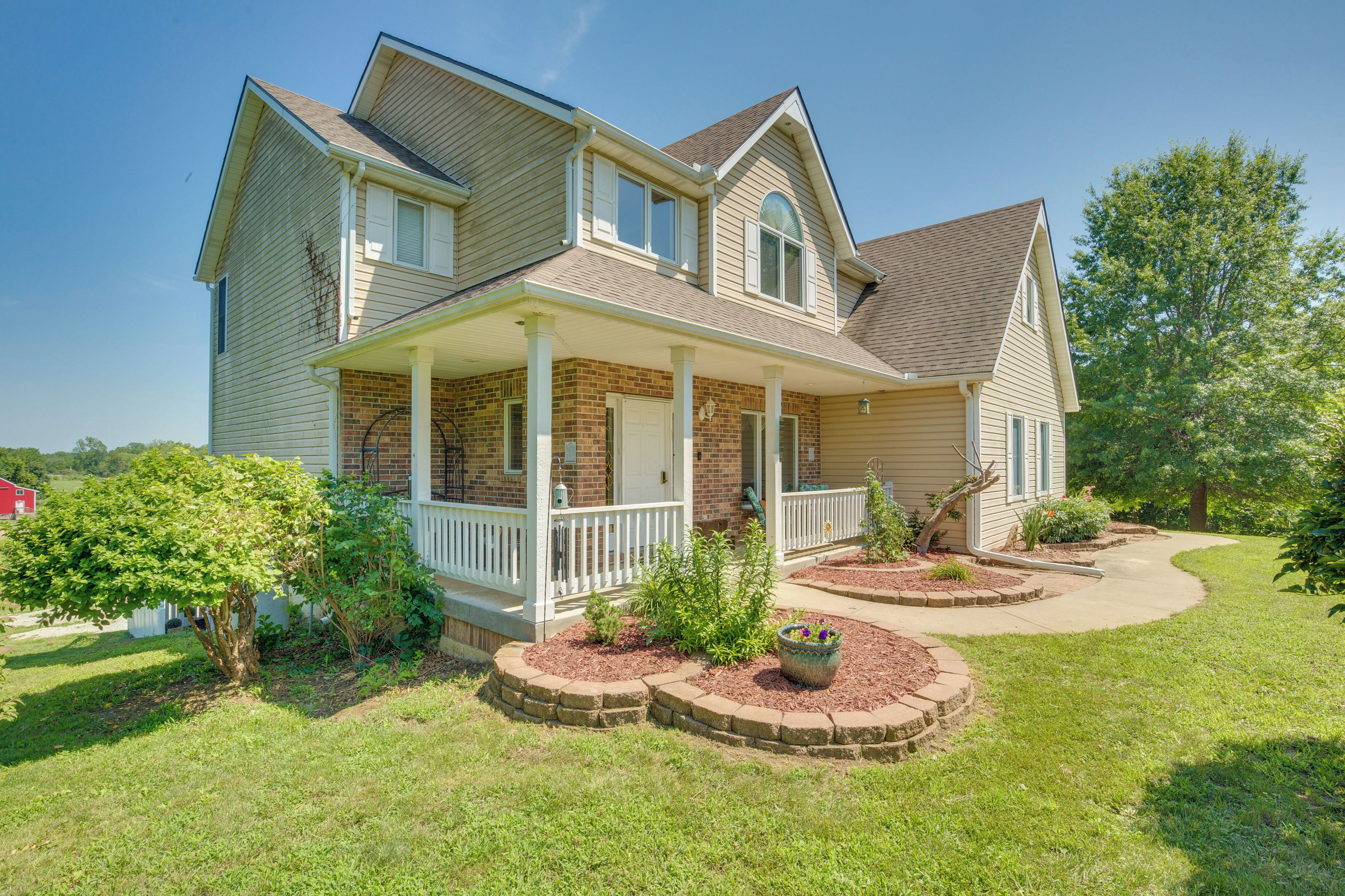 A two-story house with a front porch, brick foundation, and a landscaped yard with a tree and flower bed.