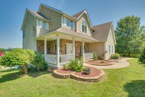A two-story house with a front porch, brick foundation, and a landscaped yard with a tree and flower bed.