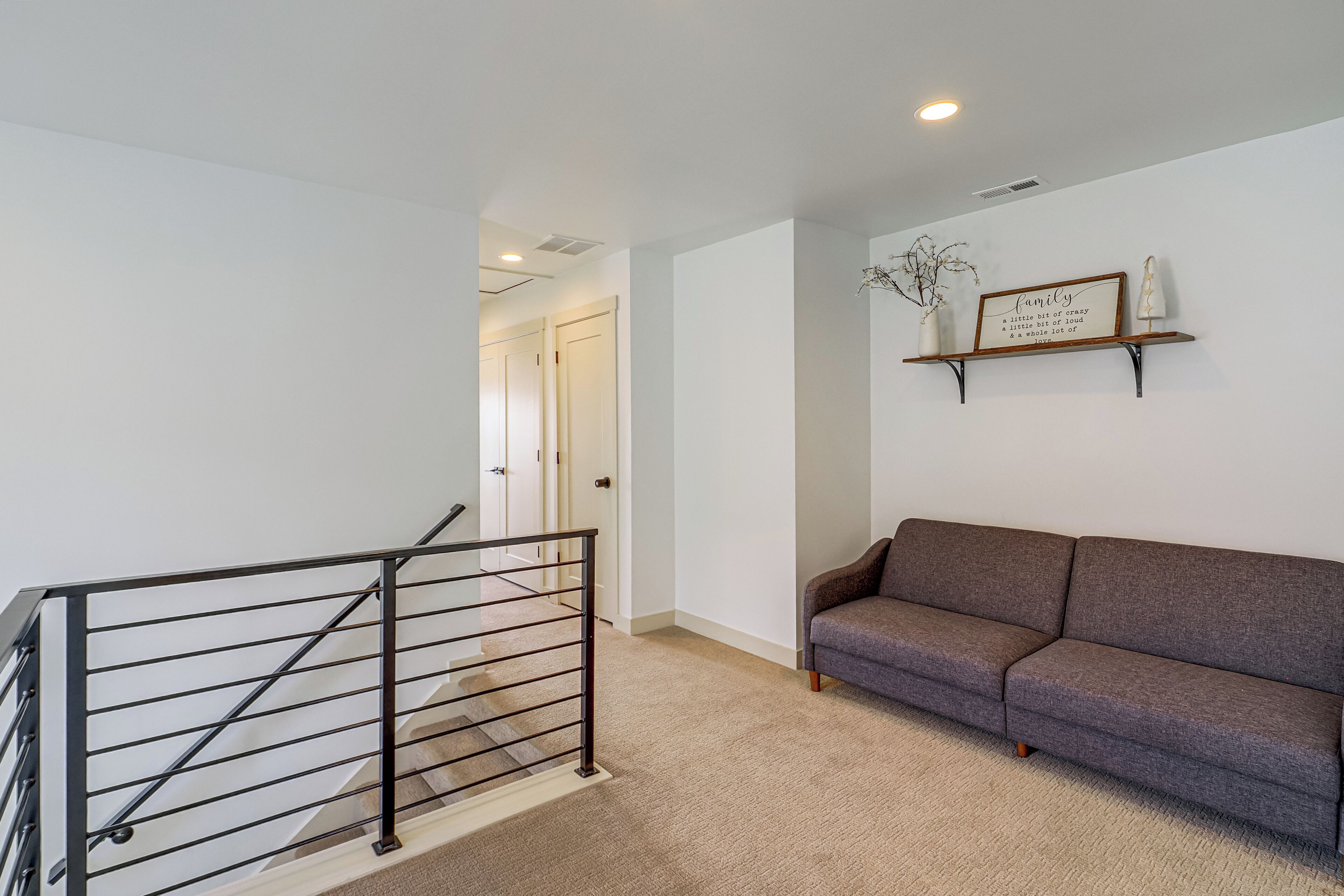 Eine Treppe mit Metallgeländer, das zu einer Zwischenebene führt, ein graues Sofa und ein an der Wand befestigtes Regal mit einem dekorativen Schild.