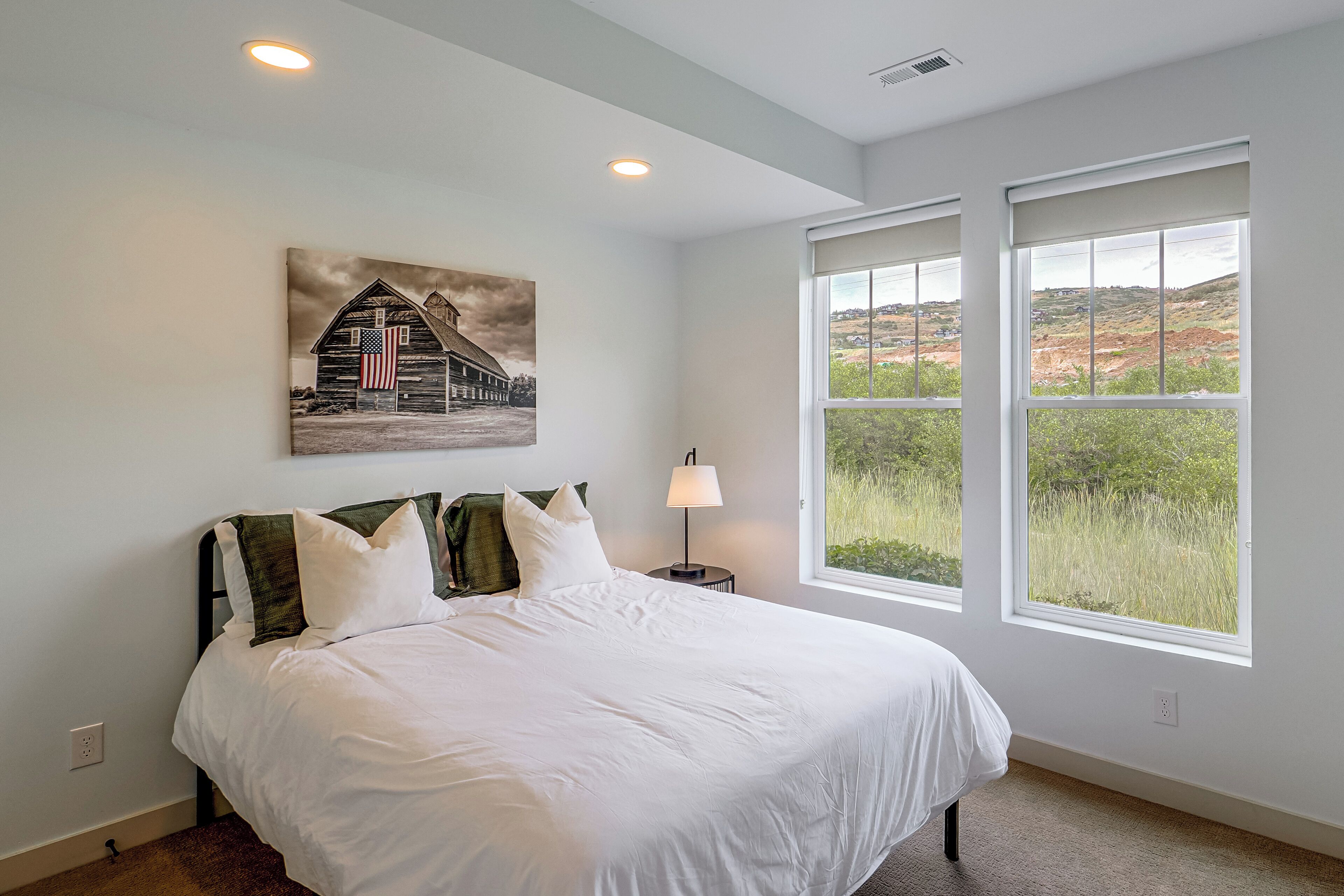 A bedroom with a large bed, two windows, a painting of a barn, and a lamp.