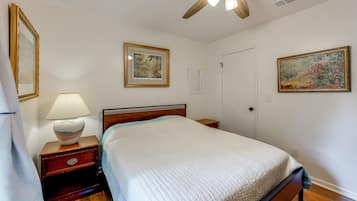 Un dormitorio con una cama, una mesita de noche, una lámpara, un ventilador de techo y dos cuadros en la pared.