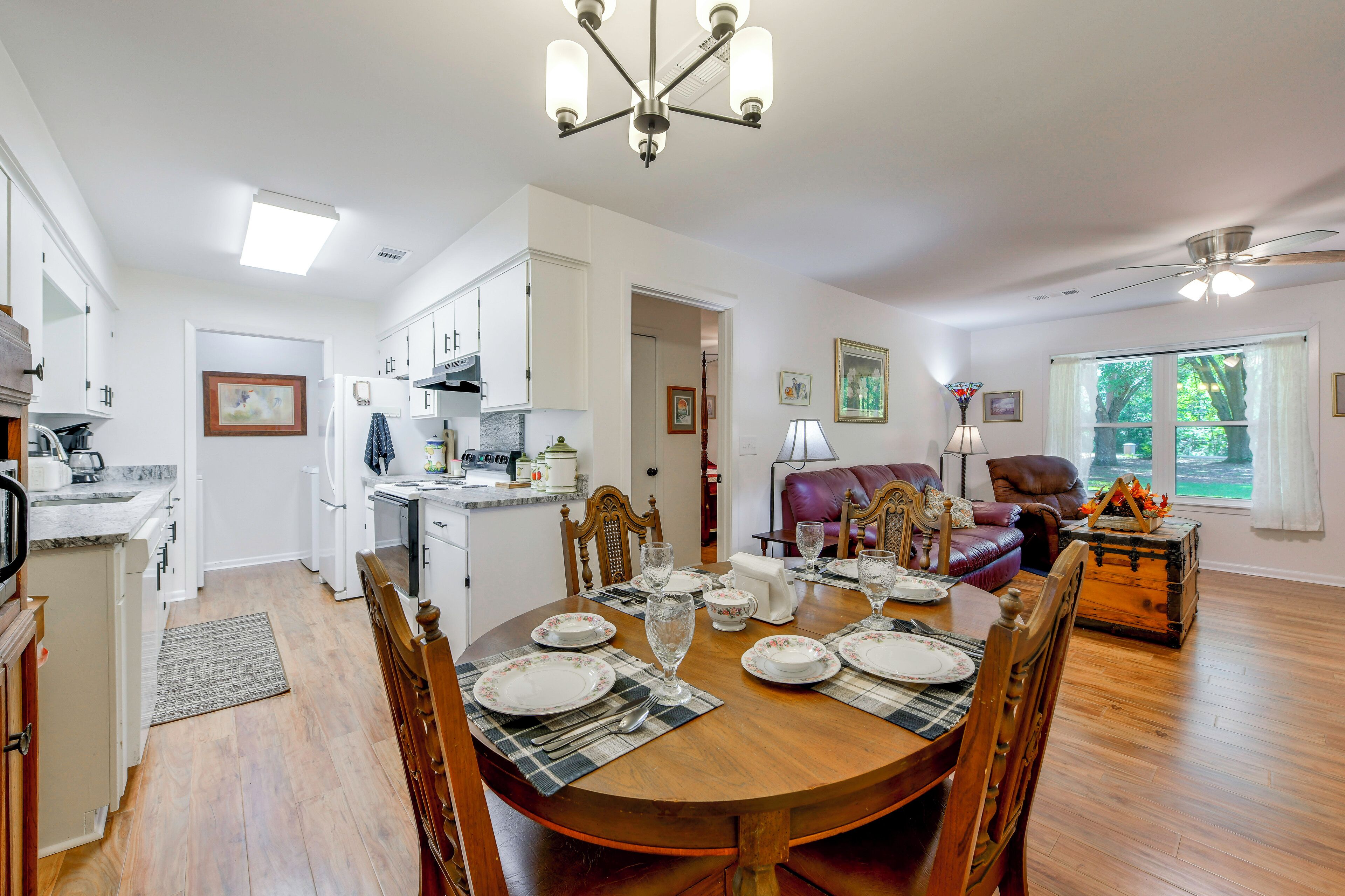 A spacious dining area with a wooden table set for four, surrounded by chairs. A kitchen with white cabinets and a refrigerator is visible in the background.