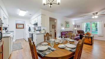 A spacious dining area with a wooden table set for four, surrounded by chairs. A kitchen with white cabinets and a refrigerator is visible in the background.