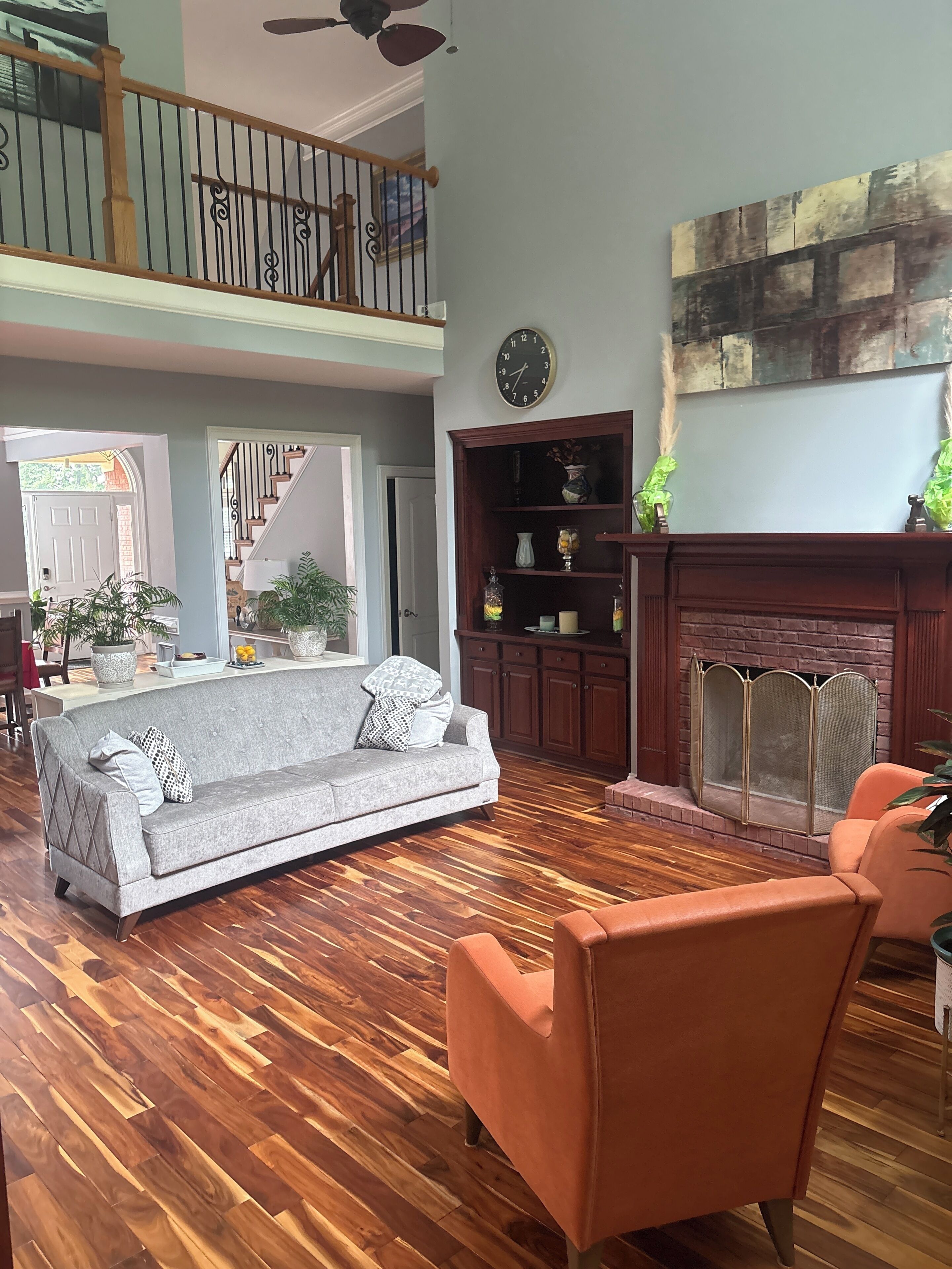 A spacious living room with a wooden floor, a grey sofa, an orange armchair, a fireplace, and a staircase leading to an upper level.