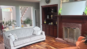 A spacious living room with a wooden floor, a grey sofa, an orange armchair, a fireplace, and a staircase leading to an upper level.