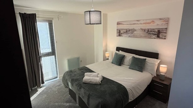 Ein Schlafzimmer mit einem Bett, Nachttischlampen, einem Fenster mit Vorhängen und einem an der Wand hängenden Kunstwerk.