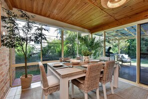 Un coin repas avec une table en bois dressée pour quatre personnes, entourée de chaises en osier, de grandes fenêtres et de plantes en pot.