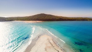 Une plage aux eaux turquoise et limpides, un rivage sablonneux et, en arrière-plan, une colline couverte d’une végétation luxuriante.