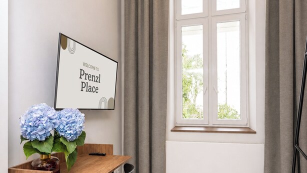 A room with a wooden desk, two chairs, a framed welcome sign, a window with curtains, and a radiator.