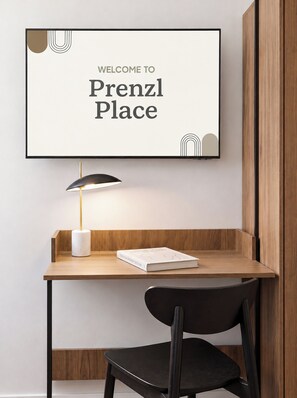 A framed welcome sign above a wooden desk with a lamp and a book, and a black chair in front. - Prenzl Place (Berlin)