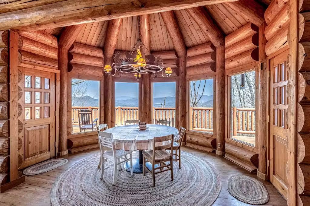 Interior de madera con una mesa redonda y sillas, ventanas amplias y vistas a montañas.