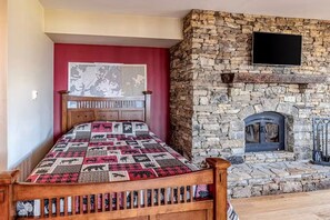 A bedroom with a stone fireplace, a bed with a patterned quilt, and a mounted television. - Two Bears | Mountain Views and Game Nights (Franklin)