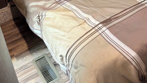 Ein Bett mit Kopfteil, ein Fenster mit Vorhängen und ein Lüftungsgitter am Boden.