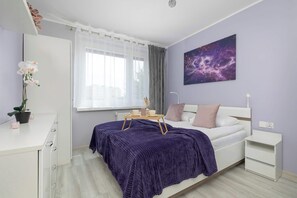 Ein Schlafzimmer mit einem Bett, Nachttischen, einem Schreibtisch, einem Fenster mit Vorhängen, einem Gemälde einer Galaxie und einer Topfpflanze.